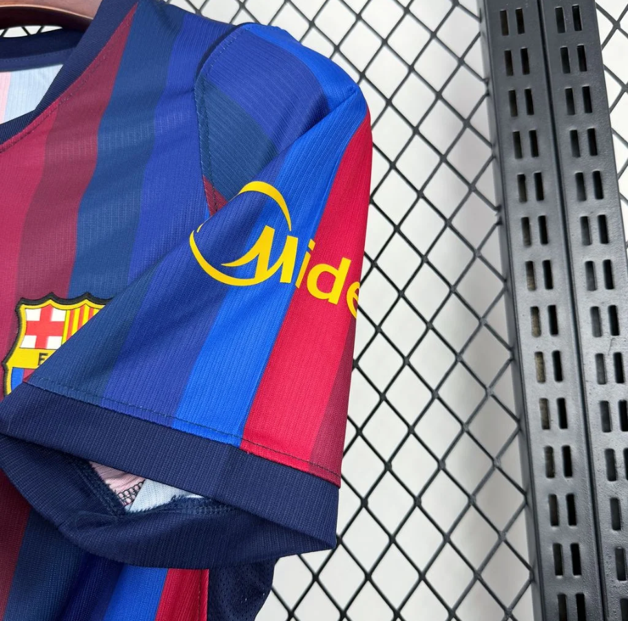 A close-up of a FC Barcelona soccer jersey with blue and red vertical stripes, featuring the yellow Midea logo on the chest and part of the FC Barcelona crest on the sleeve, hanging against a metal mesh background.