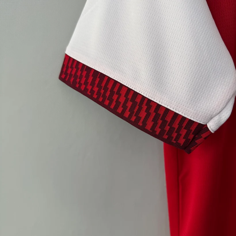 Close-up of a white athletic sleeve with a red and black geometric patterned cuff trim, shown against a light gray background.