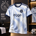 A light blue and white Ajax Amsterdam soccer jersey with navy trim, featuring the Ziggo sponsor logo and club crest, displayed in a themed interior setting.