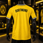 A yellow Borussia Dortmund soccer jersey with 'DORTMUND' printed in black across the back, displayed on a black mannequin in a retail store setting with a blurred BVB logo in the background.