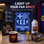 Barcelona FC LED acrylic light sign displayed on a desk alongside matching merchandise, including a white water bottle, a mug, a framed photo, a small trophy keychain, and a cube-shaped decor piece. The scene includes promotional text highlighting the bright acrylic lamp for room, desk, or shelf display.