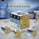 A Manchester City-themed cube puzzle set displayed on a marble surface with branded team merchandise in the background. The main product is a large cube showing the word 'MAN CITY' and multiple Manchester City logos, with three smaller cubes around it.
