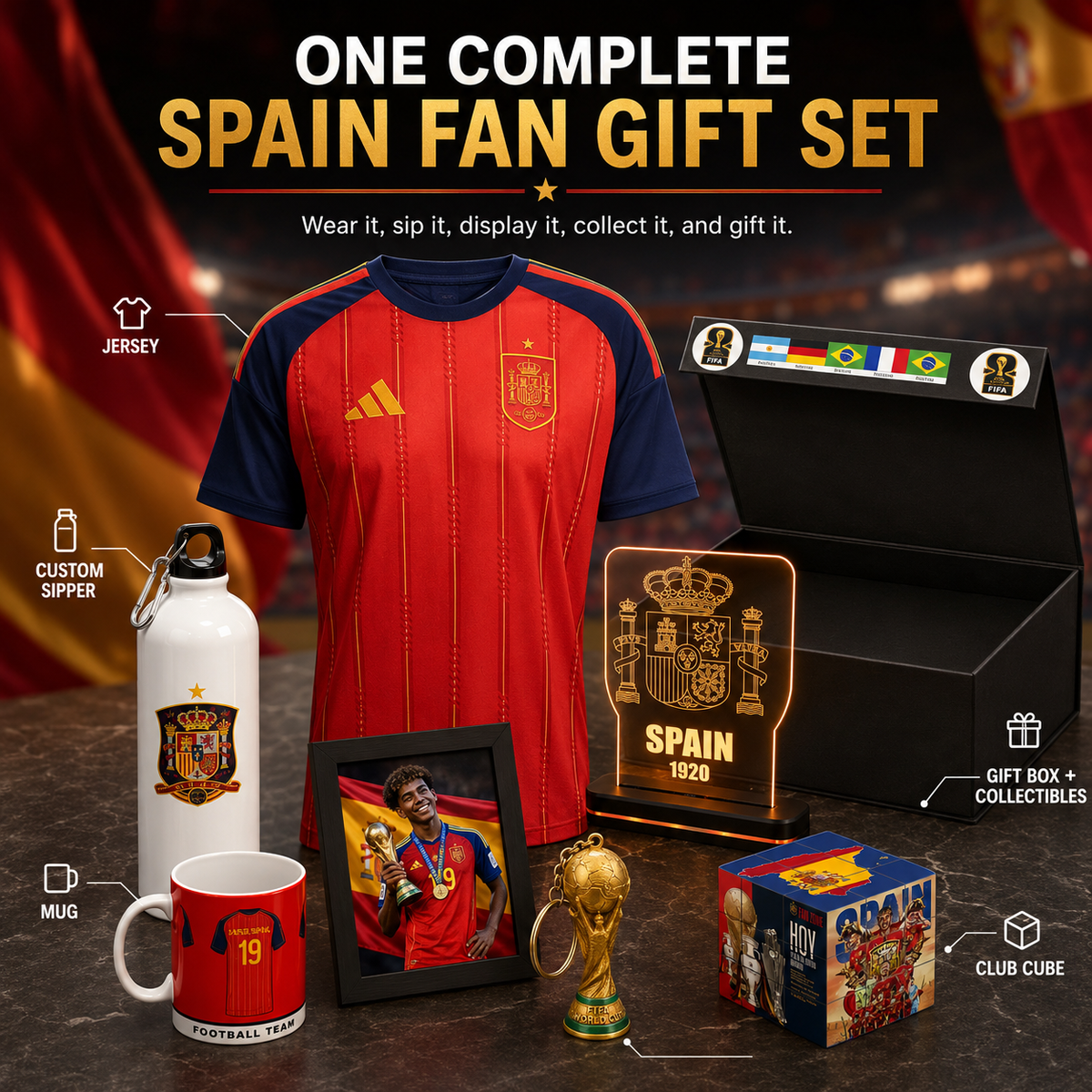 A Spain soccer fan gift set displayed on a dark tabletop with a red-and-navy jersey as the main item, plus a white water bottle, red mug, framed photo, illuminated Spain plaque, small trophy, club cube box, and black gift box. Large text at the top reads 'ONE COMPLETE SPAIN FAN GIFT SET' with callout labels for jersey, custom sipper, gift box + collectibles, mug, and club cube.