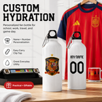 Two white personalized water bottles displayed on a wooden table, one with a Spain national team crest and the other with 'ANY NAME 00' printed on the front. A red Spain soccer jersey hangs in the background, and promotional text on the left highlights customization options.