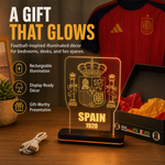 An illuminated Spain 1920 acrylic LED light display on a black base, shown on a wooden surface with a charging cable, a gift box, and a red soccer jersey in the background. Large promotional text reads 'A GIFT THAT GLOWS' with feature icons.