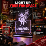 A Liverpool FC illuminated acrylic desk lamp with the club's gull logo and 'LIVERPOOL 1892' text, shown lit on a desk in a room setting with overlaid marketing callouts and surrounding football-themed decor.