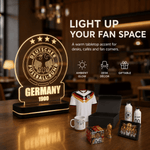 A Germany football-themed tabletop light and gift set displayed on a wooden table in a cafe-like interior. The main product is a glowing acrylic lamp with the Deutscher Fußball-Bund emblem and 'GERMANY 1900' text.