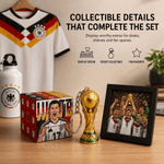 A FIFA World Cup Germany collectible set displayed on a wooden table, including a small gold FIFA World Cup trophy keychain, a framed photo of players celebrating with the trophy, a red and black patterned box, and a white Germany soccer jersey hanging in the background. Text on the image highlights collectible details of the set.