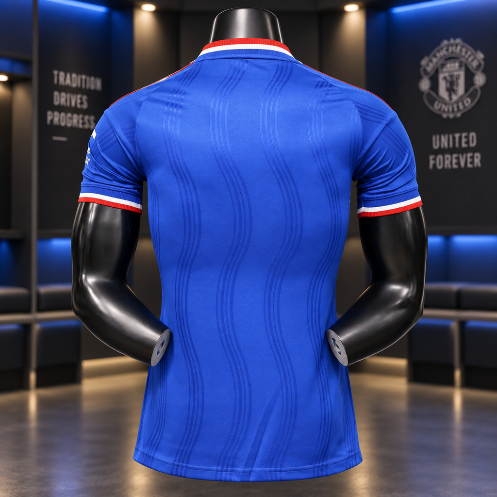 A blue short-sleeve soccer jersey shown from the back on a black mannequin torso, with red and white striped trim at the collar and sleeve cuffs. The background is a dimly lit modern interior with blue ambient lighting and Manchester United branding on the wall.