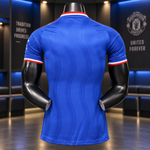 A blue short-sleeve soccer jersey shown from the back on a black mannequin torso, with red and white striped trim at the collar and sleeve cuffs. The background is a dimly lit modern interior with blue ambient lighting and Manchester United branding on the wall.