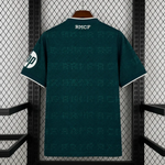 A dark teal short-sleeve soccer jersey with white trim, hanging on a wooden hanger against a black metal mesh backdrop. The back of the jersey features the white text 'RMCF' at the collar, a repeating tonal pattern, and a white circular logo on the sleeve.