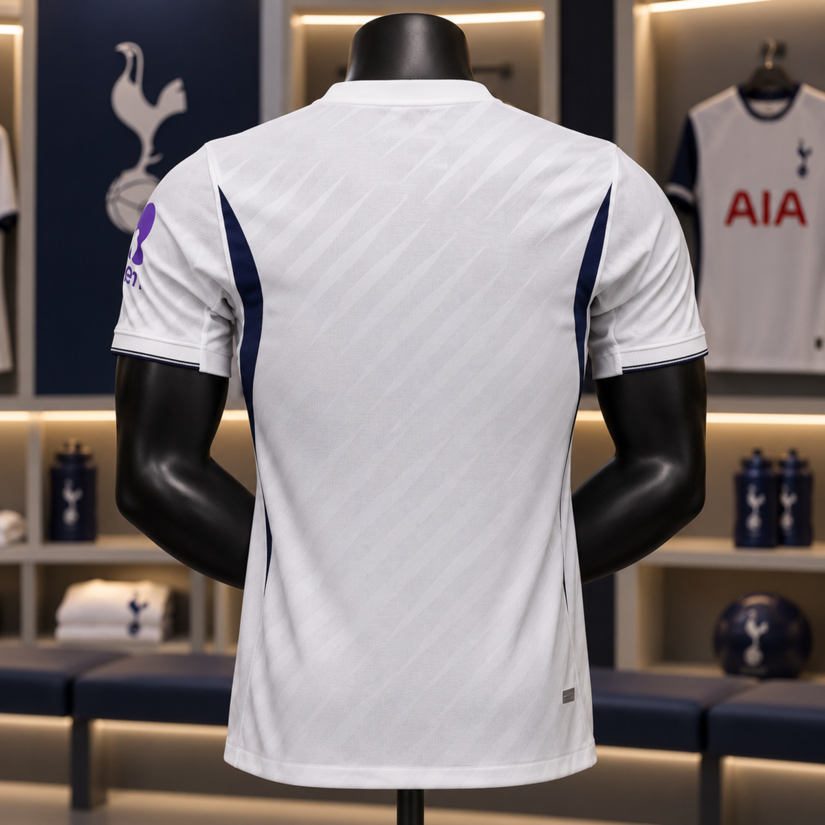 White Tottenham Hotspur football jersey shown from the back on a black mannequin, with navy side panels and a purple crest on the sleeve, set in a locker room display.