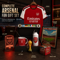 Arsenal fan gift set displayed on a dark table with a red Arsenal jersey hanging above, including a gift box, mug, water bottle, LED lamp, trophy keychain, framed stadium print, and magic cube, with text labels pointing to each item.
